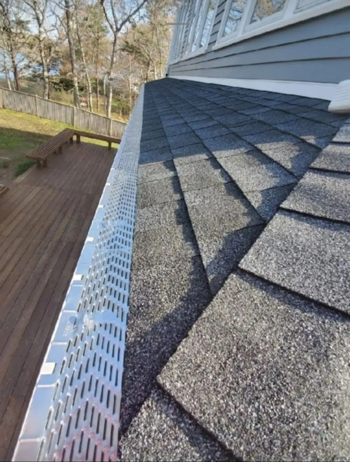 Roof shingles beside a white gutter on a house exterior, with a wooden deck and trees below.