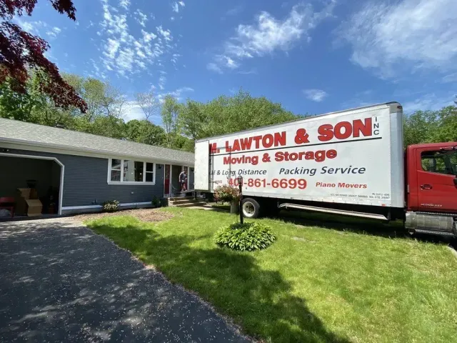 Moving truck parked in front of a grey house on a sunny day.