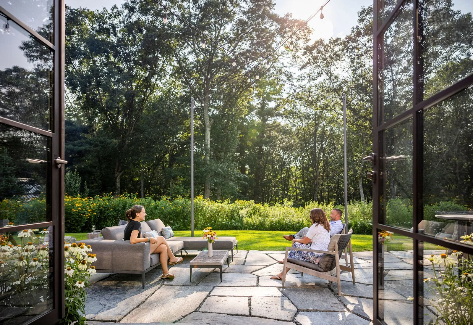Three people relax on a patio with a sofa and chairs, surrounded by greenery, flowers, and string lights, with open glass doors leading inside.