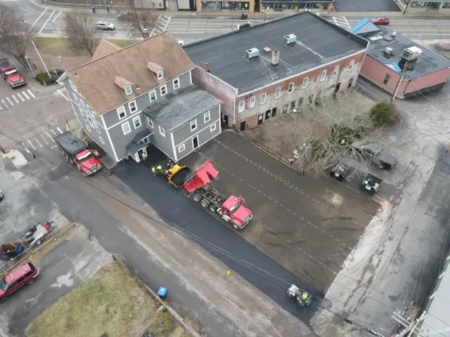 Asphalt paving in progress next to buildings; red dump trucks, yellow loader, workers.