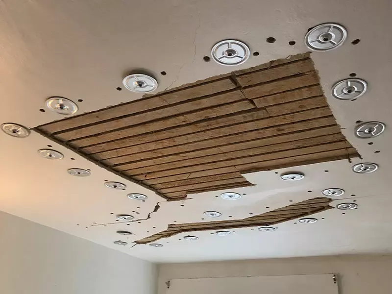 A ceiling with multiple exposed wooden beams and torn plaster, surrounded by evenly spaced recessed light fixtures.