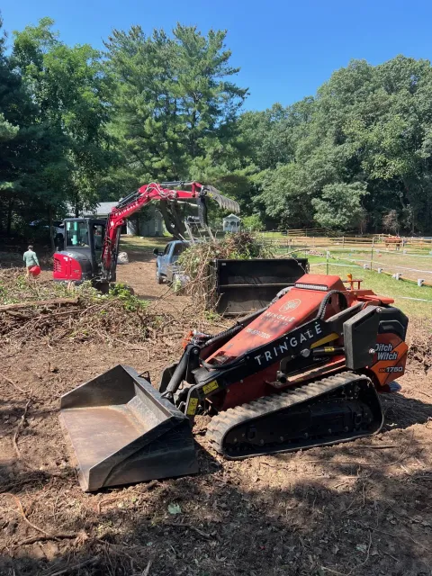 A Tringale Landscaping mini skid steer and compact excavator clearing brush and loading debris on a cleared lot