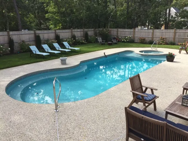 Swimming pool with lounge chairs, hot tub, and patio furniture. Green lawn, trees, and fence in the background.