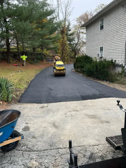 Asphalt driveway being paved with roller machine; worker in safety vest nearby.