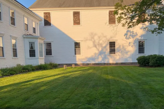 White wooden building with multiple windows and a large green lawn, featuring shadows from trees, in late afternoon sunlight.