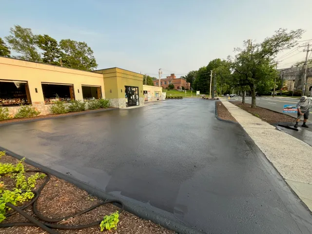 Newly paved and freshly sealed parking lot in front of a commercial building, with a worker smoothing the asphalt on the sidewalk.