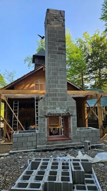 Under construction brick fireplace outside of a house, with a chimney reaching towards the sky and construction materials in the foreground.