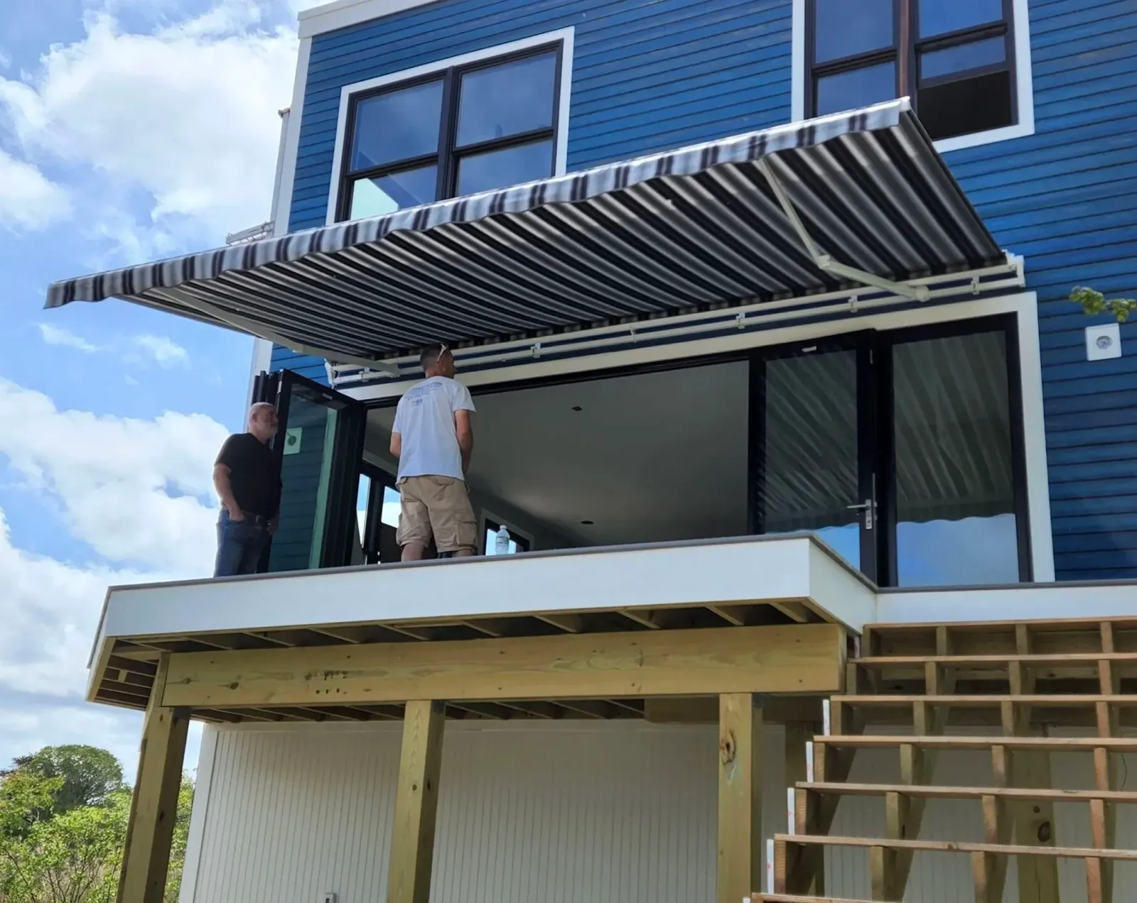 Construction workers building a new house with a blue exterior and large windows, seen from below with blue sky and clouds in the background.