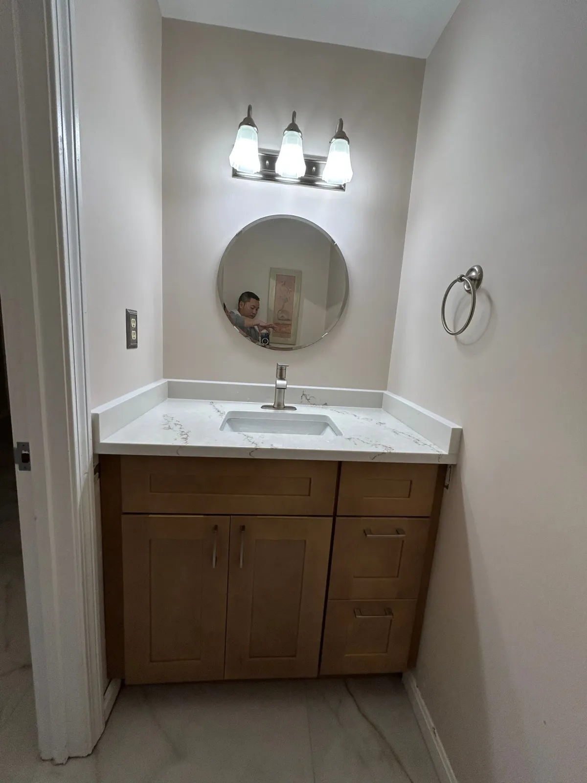 Small bathroom vanity with white sink, round mirror, and three-light sconce above a wooden cabinet.