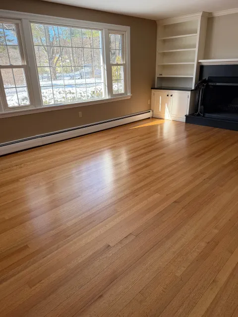 Empty living room with large window, hardwood floors, built-in shelves, fireplace, and baseboard heating.
