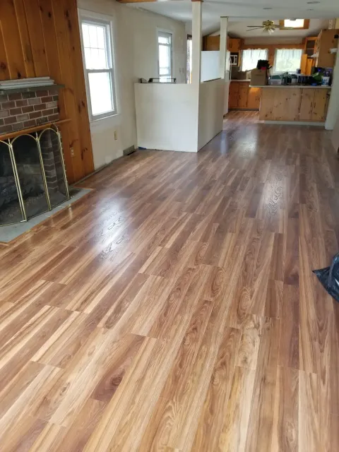 An open-concept living area with newly installed light-brown wood laminate flooring, a brick fireplace, and wood-paneled walls.