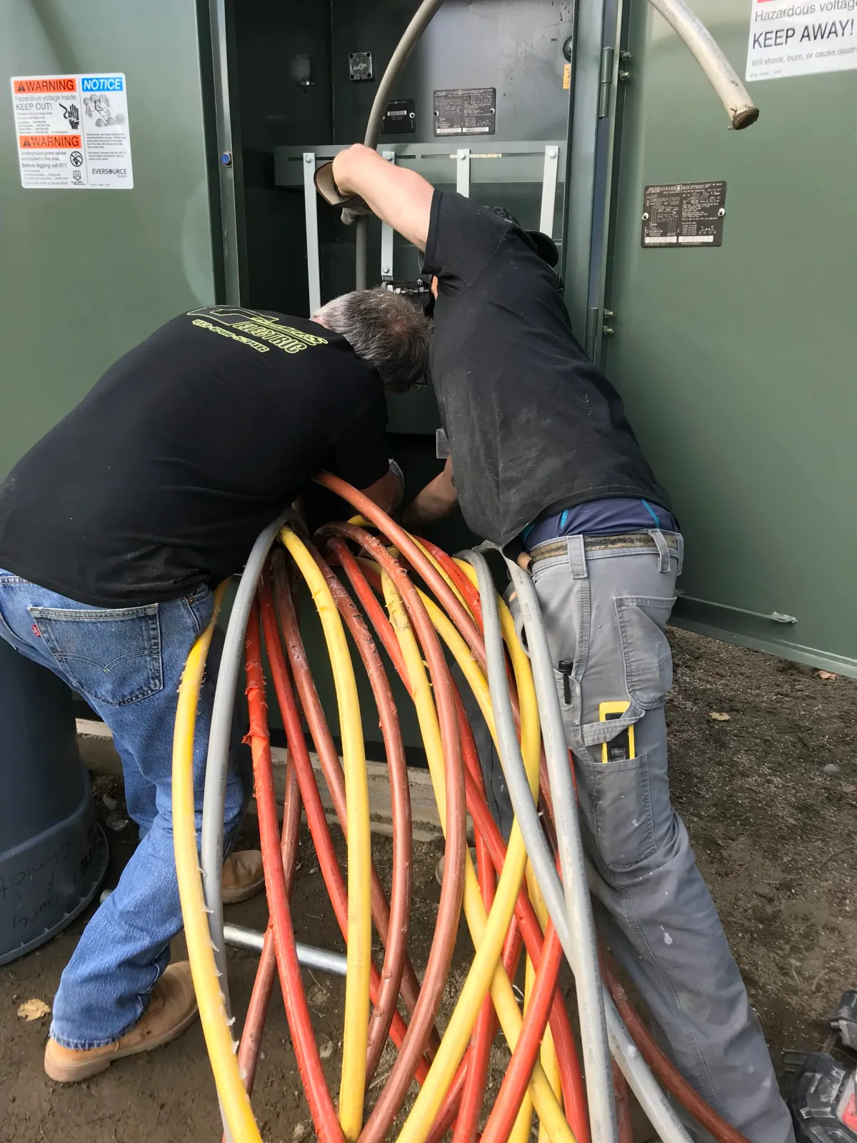 Two electricians working on an electrical panel with colorful insulated cables, wearing work clothes and tools, outside near ground level.