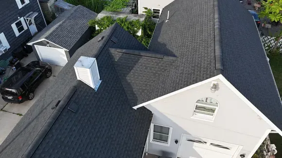 Aerial view of a house roof with dark shingles, chimneys, and neighboring homes and cars nearby