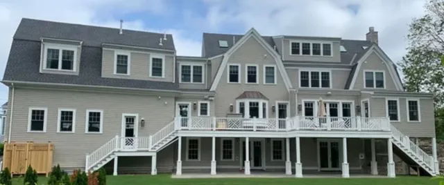 Large multi-story house with beige siding, multiple windows, a deck, and stairs leading to the yard, under a partly cloudy sky.