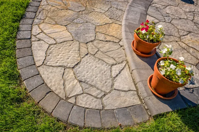 Stone paved patio with two terracotta pots containing white and pink flowers, bordered by green grass.