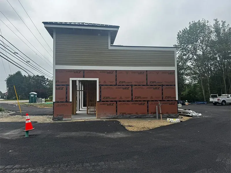Building under construction, brown siding, open doorway, green roof section, black asphalt, outdoor setting.
