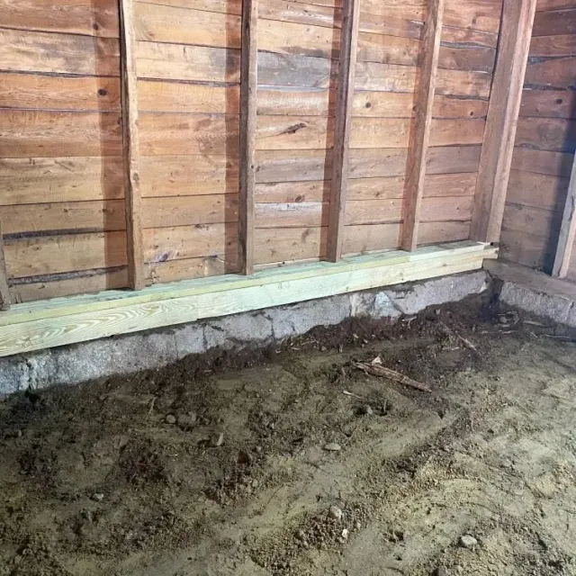 Bare dirt floor and wooden wall framing in an unfinished interior with a concrete foundation base