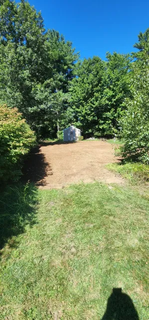 A small white shed surrounded by trees and a patch of bare, tilled soil, with some grass in the foreground under a clear blue sky.