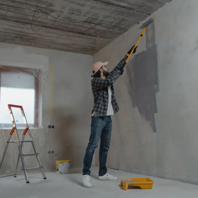 Painter rolling grey paint onto a bare wall beside a step ladder and paint tray