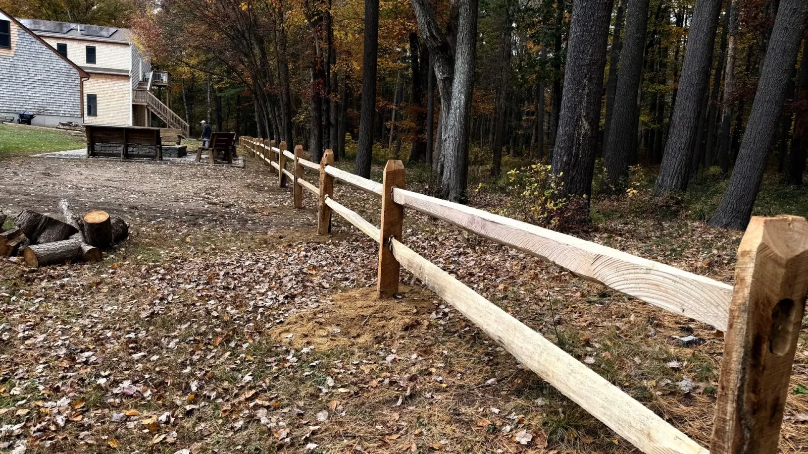Wooden split-rail fence in a wooded area with a building in the background. Autumn leaves on the ground.