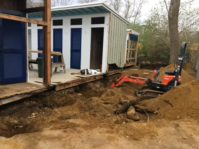 Excavator digging under a small, white and blue building with blue shutters; construction in progress.