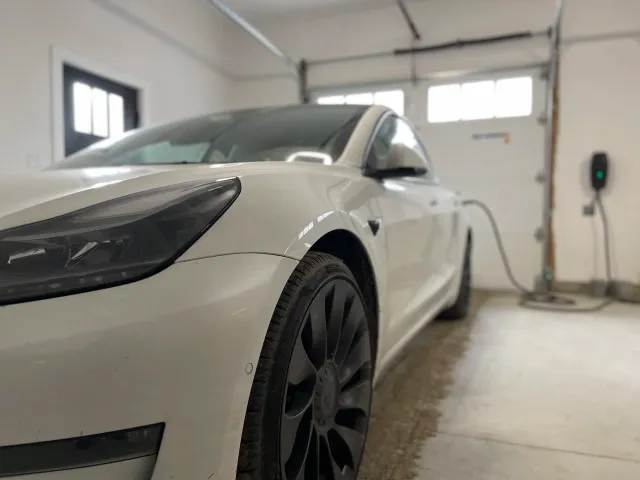 Close up of the front of an electric car that is connected by cable to a charger in the wall of a garage