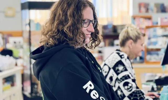 A ReSOURCE Employee smiles while working at a counter in what appears to be a ReSOURCE store or library. Another person in a black-and-white sweater is in the background, focused on a computer, creating an inviting Home Page atmosphere for visitors.