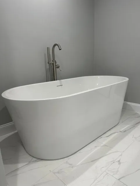 White oval bathtub with tall chrome faucet against gray wall, on marble-look floor.
