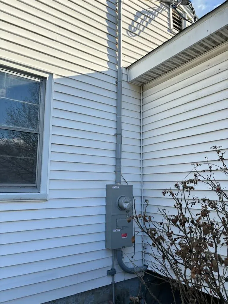 Exterior of a house with white vinyl siding, showing an electric meter box and conduit pipe. A leafless shrub is in the foreground.