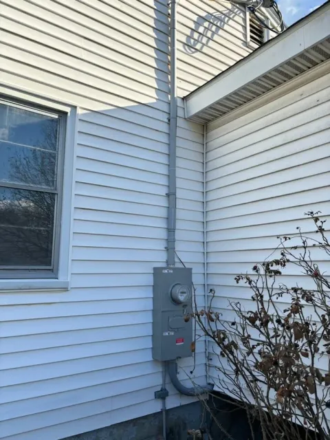 Exterior of a house with white vinyl siding, showing an electric meter box and conduit pipe. A leafless shrub is in the foreground.