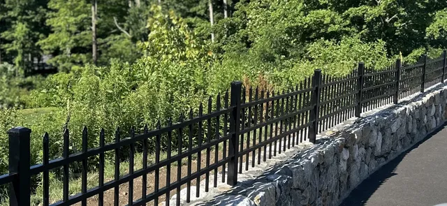 Black aluminum spear-top fence mounted along a mortared stone wall beside a paved driveway with greenery behind