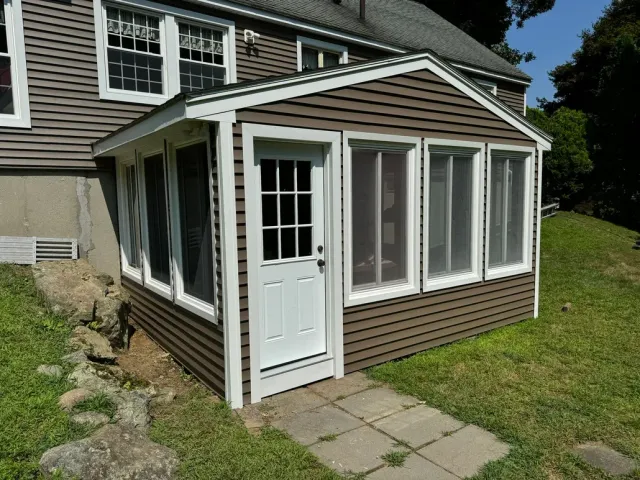 Small brown sunroom addition with white trim and door beside a house, with patio stones and grass.