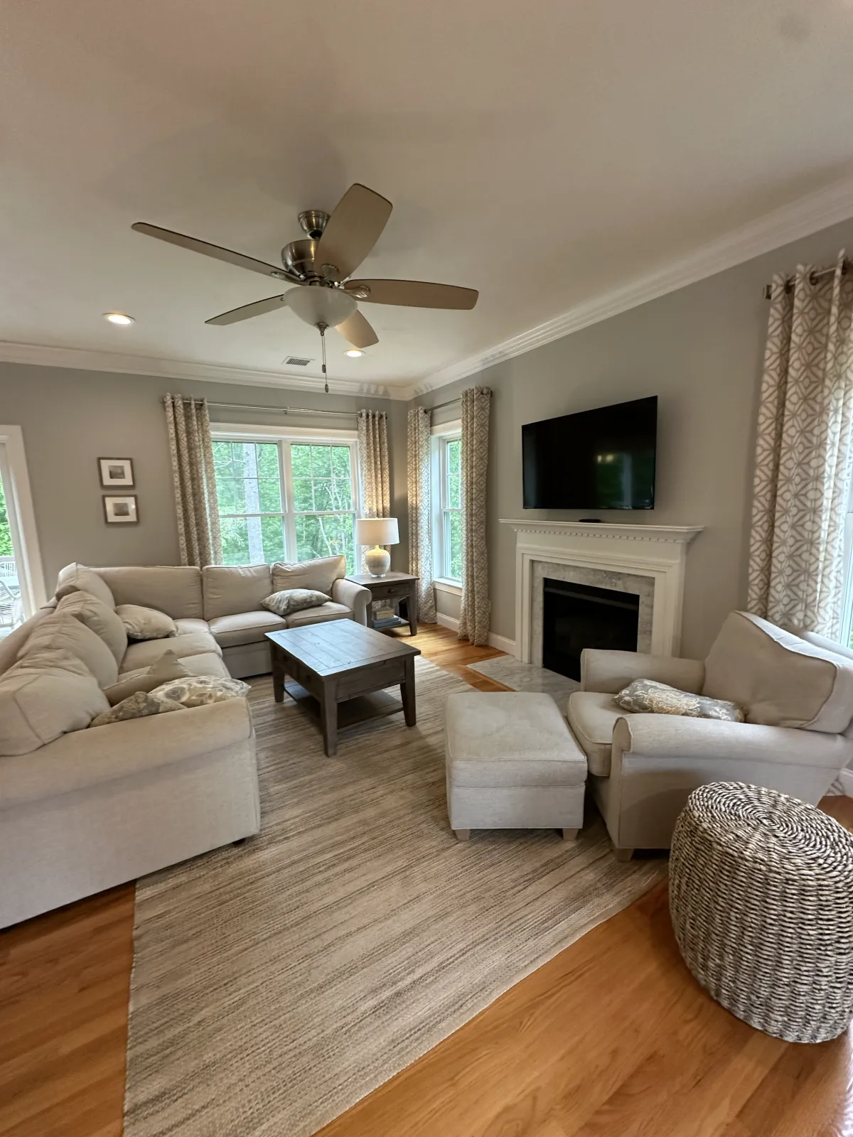 Living room with beige sofas, a wooden coffee table, a fireplace, a wall-mounted TV, windows with patterned curtains, a ceiling fan, and hardwood flooring.
