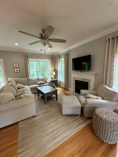 Living room with beige sofas, a wooden coffee table, a fireplace, a wall-mounted TV, windows with patterned curtains, a ceiling fan, and hardwood flooring.