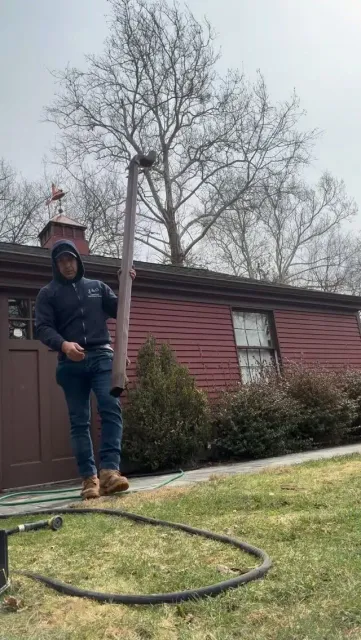 Person in hoodie standing on lawn with a hose in front of a red house and leafless tree