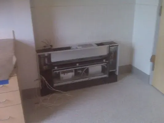 A partially disassembled white wall-mounted HVAC cabinet in a modern office or institutional room.