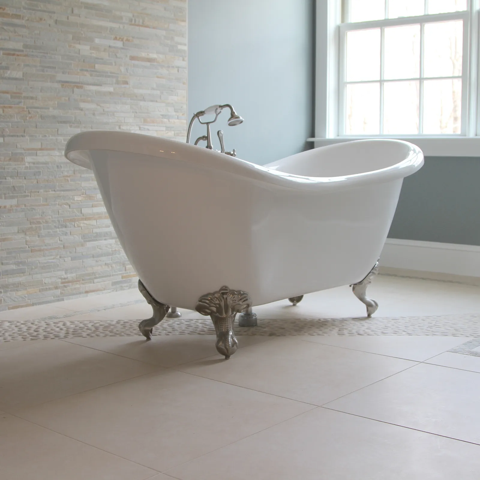 White clawfoot bathtub with silver feet on beige tile floor, stone accent wall, and pebble tile border.