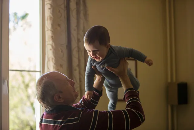 An elderly man lifting a young child, who is smiling and reaching out, indoors with a window and curtains in the background.