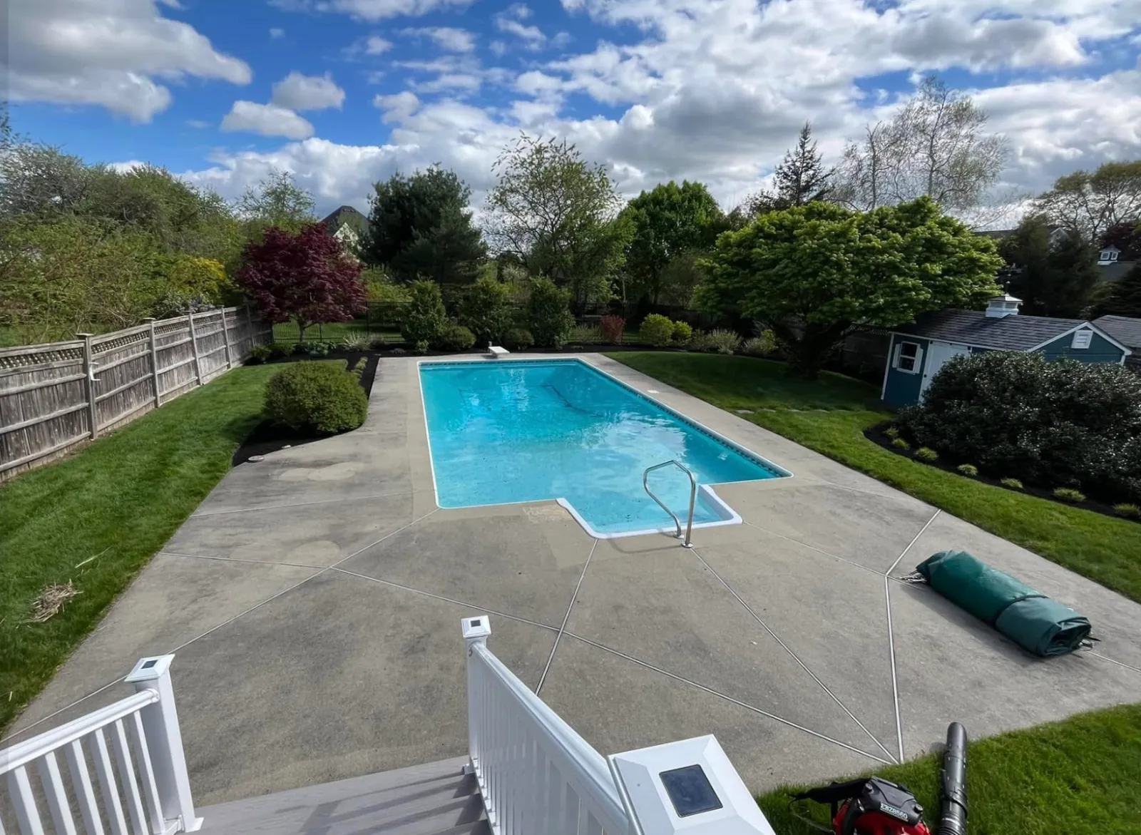 A backyard swimming pool surrounded by a concrete deck, with a garden and trees, and a fence separating it from neighboring houses. The sky is partly cloudy.