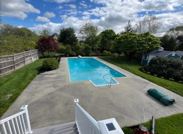 A backyard swimming pool surrounded by a concrete deck, with a garden and trees, and a fence separating it from neighboring houses. The sky is partly cloudy.