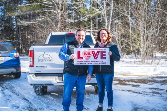 Alt Text: Rob Wilkes and Kelsey Cole of Colby & Gale holding a "Fuel Your Love" sign to promote community warmth and support in Midcoast Maine.