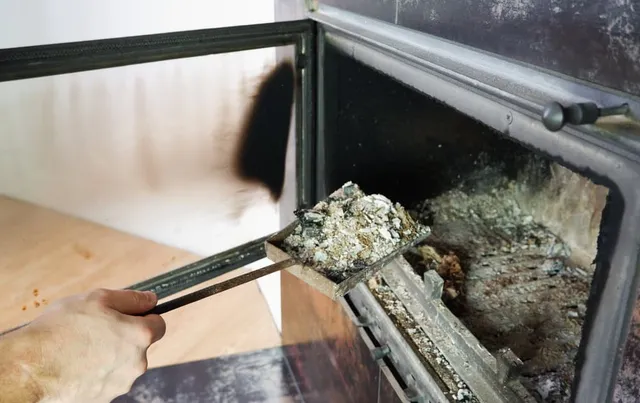 A person uses a metal tool to scoop out ashes from a wood-burning stove, with the stove door open and soot visible on the glass.