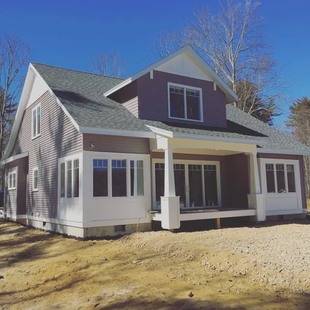 Newly built house with purple siding, white trim, large windows, a covered porch, and a gray shingled roof, on a construction site with dirt ground and leafless trees in the background.