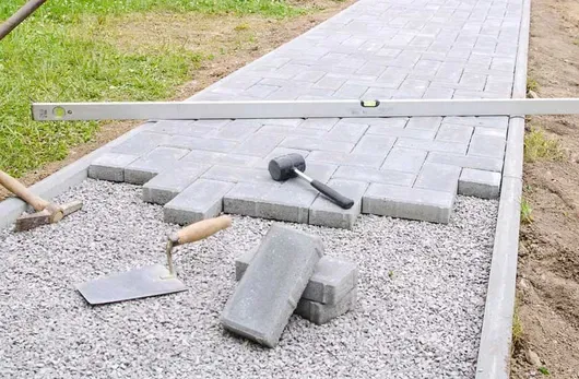 Broken paving stones and tools scattered on a gravel path beside a partially repaired walkway.