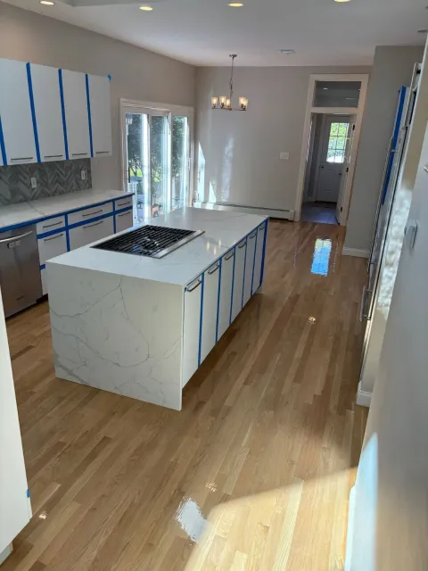 kitchen with all white cabinets that have been taped off with blue painters tape, a marble waterfall island, and shiny hardwood floors that have just been installed