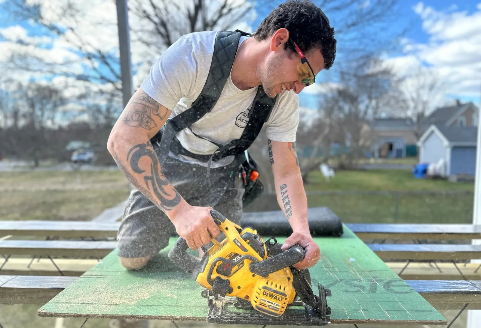 A man with tattoos wearing safety glasses and a grey t-shirt uses a yellow DeWalt cordless saw, working on a green wooden surface outdoors with a blue sky and houses in the background.