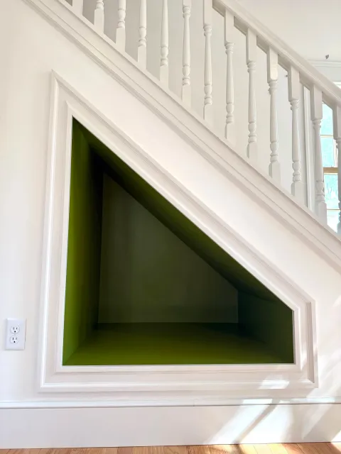 A built-in corner nook with green painted interior, framed by white molding, underneath a staircase with white spindles and railing.