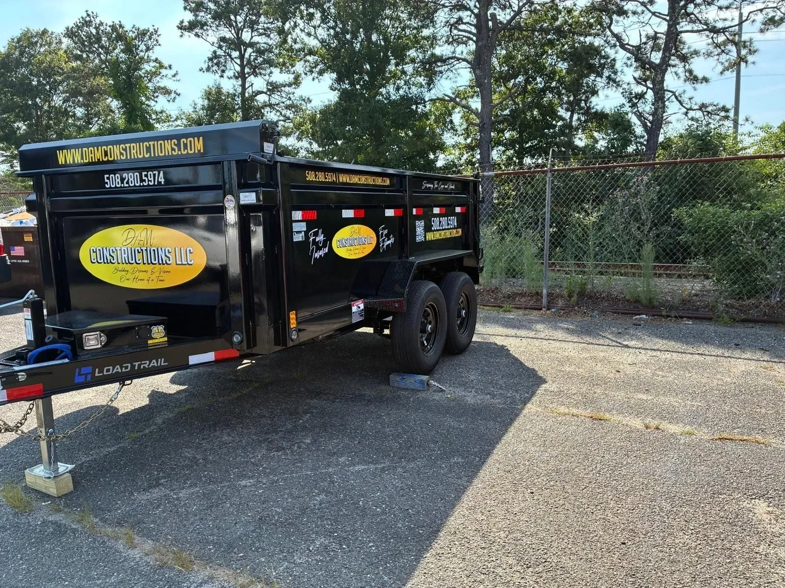 Black utility trailer parked on gravel, with yellow logos and a chain-link fence in the background
