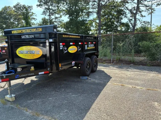 Black utility trailer parked on gravel, with yellow logos and a chain-link fence in the background