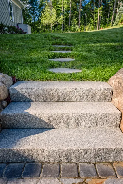 Stone steps leading up to a grassy hill with a path of stepping stones in a garden, surrounded by trees.